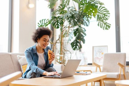 Photo for Happy attractive African American woman with curly hair sitting on comfortable sofa, eating croissant, using laptop, working in cozy cafe. Concept of remote job, lunch, online technology - Royalty Free Image