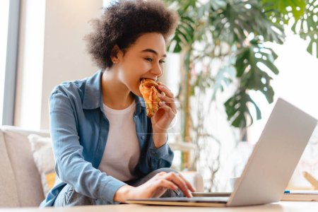Photo for Authentic, beautiful African American woman, freelancer with curly hair sitting on comfortable sofa, eating croissant, using laptop, working in co-working space. Concept of lunch, online technology - Royalty Free Image