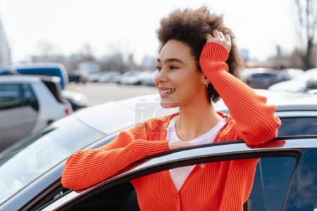 Foto de Retrato de una auténtica mujer afroamericana sonriente con el pelo rizado, de pie cerca del coche, mirando hacia otro lado. Concepto de seguro de coche, vehículo - Imagen libre de derechos