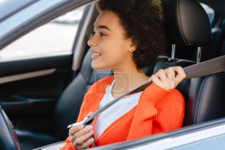 Foto de Hermosa, sonriente, joven mujer afroamericana, conductor con el pelo rizado se abrocha el cinturón de seguridad, sentado en el salón de automóviles. Concepto de seguro de automóvil - Imagen libre de derechos