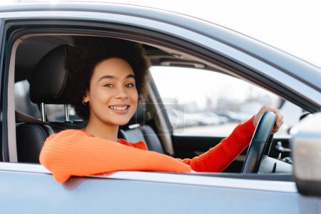 Foto de Atractiva, sonriente mujer afroamericana conduciendo coche, mirando a la cámara. Concepto de transporte, vehículo - Imagen libre de derechos