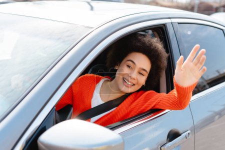 Foto de Mujer alegre, hermosa, afroamericana conduciendo coche, saludando, mirando hacia otro lado, saludando. Concepto de transporte, vehículo - Imagen libre de derechos