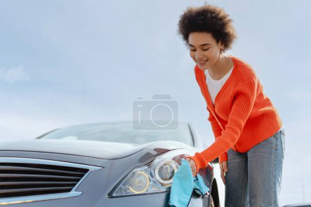 Retrato de una joven afroamericana sonriente limpiando el coche con tela de microfibra en la estación de autoservicio. Concepto de lavado de coches, limpieza, viajes