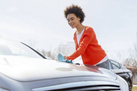 Portrait of happy, beautiful African American woman wiping down car with microfiber cloth. Concept of car washing, cleanliness, travel