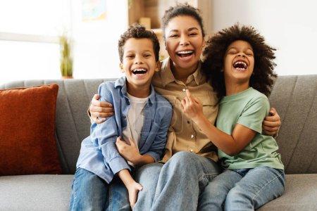 Téléchargez les photos : Famille afro-américaine souriante, mère étreignant son péché et sa fille tout en s'amusant à la maison, assise sur le canapé. Concept d'enfance, maternité, famille, plaisir - en image libre de droit