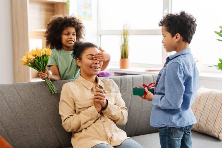 Photo for Beautiful African American family, smiling children giving gift and bouquet of yellow tulips to excited mother, sitting at home on cozy sofa. Concept of Mothers Day, celebration - Royalty Free Image
