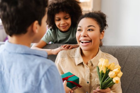 Téléchargez les photos : Surprise femme afro-américaine recevant des cadeaux de ses enfants, tenant un bouquet de fleurs et boîte cadeau à la maison. Concept de fête des mères, célébration - en image libre de droit