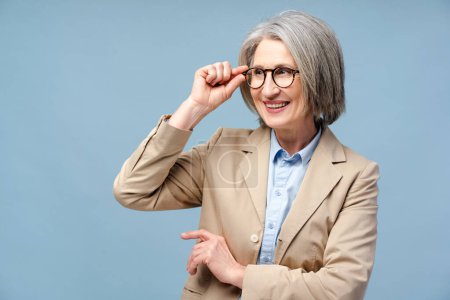 Téléchargez les photos : Portrait de femme âgée souriante portant des lunettes élégantes isolées sur fond bleu, concept de vision. Jolie femme d'affaires aux cheveux gris heureux, gestionnaire confiant détournant les yeux - en image libre de droit