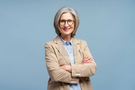Smiling senior business woman wearing eyeglasses looking at camera, posing arms crossed isolated on blue background. Portrait of confident gray haired politician. Successful business, career