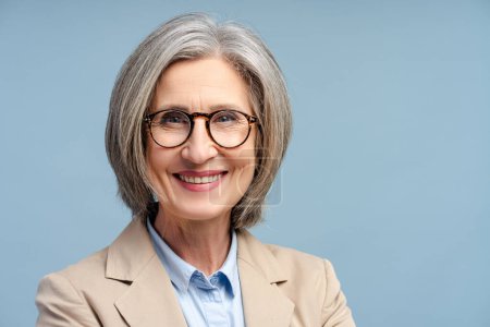 Smiling senior business woman, manager, financier wearing eyeglasses looking at camera isolated on blue background. Portrait of confident gray haired politician. Successful business, career