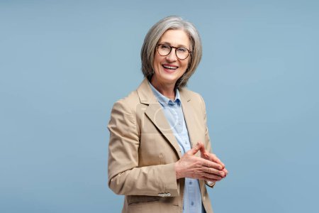 Smiling senior business woman wearing eyeglasses looking at camera, posing isolated on blue background. Portrait of confident gray haired politician. Successful business, career