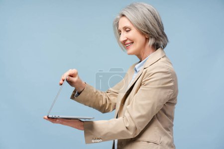 Téléchargez les photos : Sourire femme âgée aux cheveux gris ordinateur portable d'ouverture de travail en ligne isolé sur fond bleu. Portrait confiant 60 ans auteur réussi regardant l'écran. Concept technologique - en image libre de droit