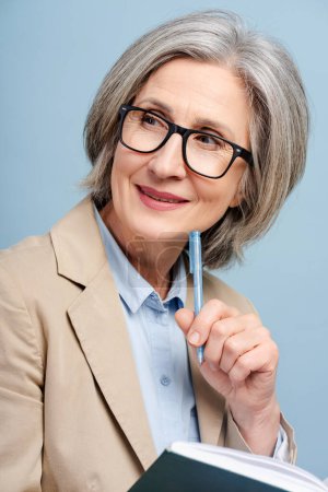 Photo for Portrait pensive senior woman, writer taking notes, holding book, brainstorming isolated on blue background. Attractive gray haired worker, manager looking away planning startup - Royalty Free Image