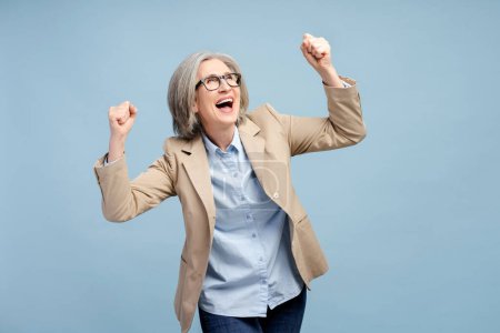 Photo for Portrait of excited senior woman in suit making winner gesture rejoicing isolated on blue background. Victory concept - Royalty Free Image
