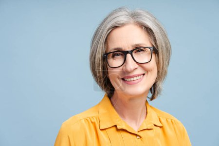 Attractive smiling senior woman, happy grandmother wearing eyeglasses and casual yellow blouse, looking at camera standing isolated on yellow background. Retired concept