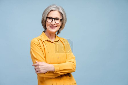 Attractive smiling senior woman, happy grandmother wearing eyeglasses and yellow blouse, looking at camera, posing arms crossed isolated on yellow background. Retired concept