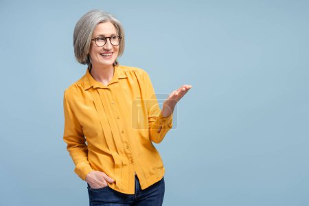 Foto de Mujer mayor sonriente con gafas elegantes apuntando con la palma en el espacio vacío, aislada sobre fondo azul. Moderno profesor de pelo gris mirando hacia otro lado, presentación - Imagen libre de derechos