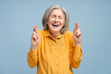 Smiling senior woman wearing casual shirt with crossed fingers standing isolated on blue background with closed eyes. Concept of wish something