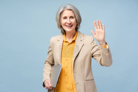 Photo for Portrait smiling senior woman, manager, worker holding laptop looking at camera and greeting hand isolated on blue background. Business concept - Royalty Free Image