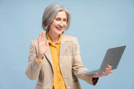 Téléchargez les photos : Heureuse femme âgée aux cheveux gris agitant la main à l'ordinateur portable travaillant en ligne isolé sur fond bleu. Portrait confiant réussi 60 ans dame ayant appel vidéo. Concept technologique - en image libre de droit