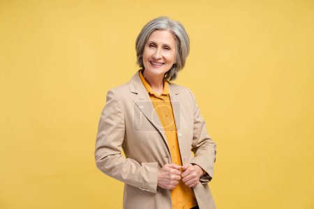 Smiling senior business woman, manager, financier looking at camera isolated on yellow background. Portrait of confident gray haired politician. Successful business, career