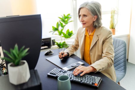 Photo for Focused smiling senior woman, graphic designer, editor, using tablet working in modern office. Technology, successful business concept - Royalty Free Image