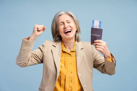 Glücklich aufgeregte grauhaarige Seniorin mit Pass und Fahrkarte, jubelnd auf blauem Grund, eine Bordkarte in der Hand, genießt den bevorstehenden Flug. Pensionist auf Auslandsreise 