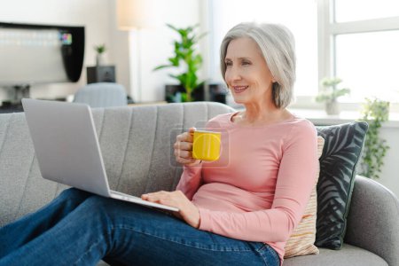 Foto de Mujer mayor sonriente usando un ordenador portátil, sosteniendo una taza de café, sentada en un cómodo sofá en casa. Mujer de negocios de pelo gris trabajando desde casa - Imagen libre de derechos