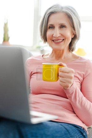 Téléchargez les photos : Vue verticale d'une femme âgée heureuse utilisant un ordinateur portable, tenant une tasse de café, assise sur un canapé confortable à la maison. Femme d'affaires aux cheveux gris travaillant à domicile - en image libre de droit