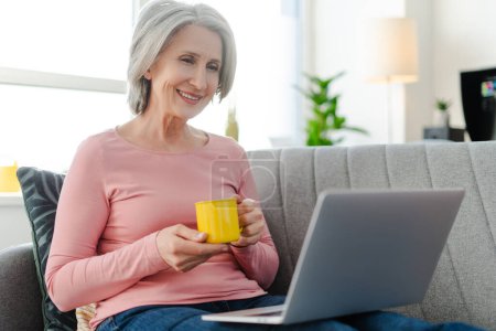 Téléchargez les photos : Femme âgée souriante utilisant un ordinateur portable, tenant une tasse de café, assise sur un canapé confortable à la maison. Femme d'affaires aux cheveux gris travaillant à domicile - en image libre de droit