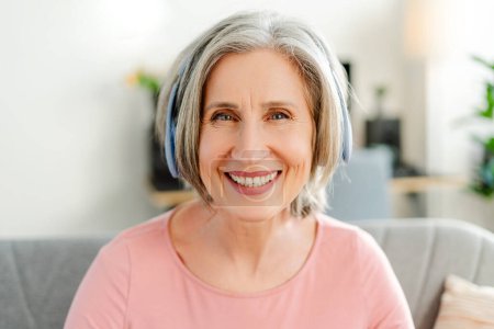 Photo for Overjoyed senior woman listening music in wireless headphones, looking at camera sitting on sofa at home. Technology, positive lifestyle concept - Royalty Free Image