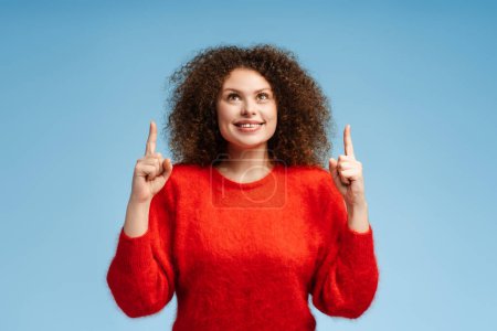 Photo for Smiling attractive woman with curly hair wearing stylish red sweater pointing fingers up, looking up at copy space, standing isolated on blue background. Advertisement concept - Royalty Free Image