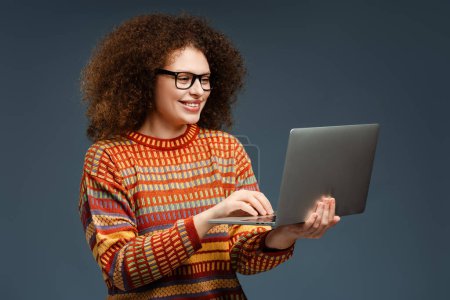Téléchargez les photos : Portrait de belle femme souriante aux cheveux bouclés à l'aide d'un ordinateur portable portant un pull confortable élégant et des lunettes tapant sur le clavier isolé sur fond gris. Concept technologique - en image libre de droit