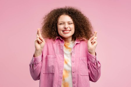 Foto de Smiling, excited, beautiful woman with curly hair, hipster with crossed fingers and closed eyes standing isolated on pink background. Concept of hope - Imagen libre de derechos