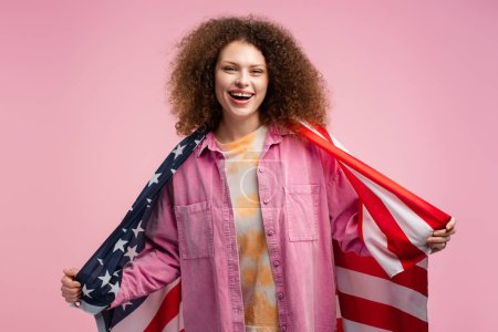 Téléchargez les photos : Joyeux jeune femme aux cheveux bouclés, patriote tenant le drapeau américain, regardant la caméra, posant isolé sur fond rose. Concept d'élection américaine, soutien, fête de l'indépendance - en image libre de droit