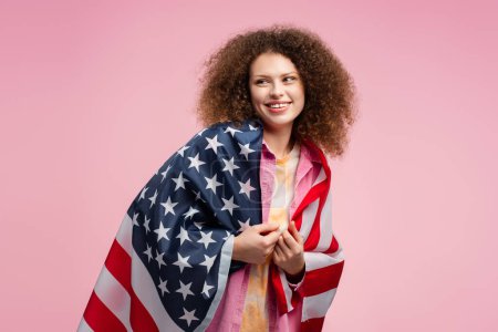 Téléchargez les photos : Joyeux jeune femme aux cheveux bouclés, patriote tenant le drapeau américain, regardant ailleurs, posant isolé sur fond rose. Concept d'élection américaine, soutien, fête de l'indépendance - en image libre de droit