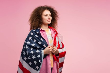 Téléchargez les photos : Belle femme attrayante avec les cheveux bouclés, supporter tenant drapeau américain, regardant loin sur l'espace de copie, debout isolé sur fond rose. Concept de l'élection présidentielle américaine de 2024 - en image libre de droit