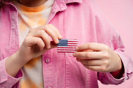 Young woman holding American flag icon, close up, selective focus on hands isolated on pink background. USA 2024 election concept, American symbol