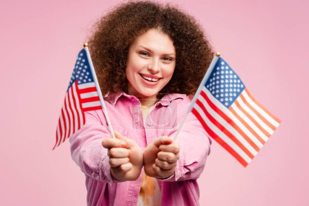 Téléchargez les photos : Jeune femme mignonne excitée avec les cheveux bouclés, supporter, patriote tenant drapeau américain regardant la caméra, debout isolé sur fond rose. Concept de l'élection présidentielle américaine de 2024 - en image libre de droit