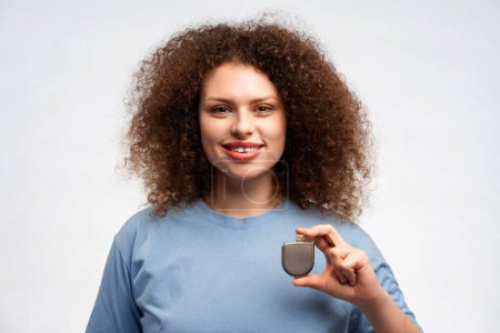 Photo for Portrait of attractive woman with curly hair in blue t shirt holding pacemaker in hand, looking at camera isolated on white background. Health care, treatment concept - Royalty Free Image