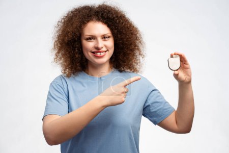 Photo for Attractive smiling young woman with curly hair holding pacemaker pointing finger at it looking at camera standing isolated on white background. Concept of treatment, health care - Royalty Free Image