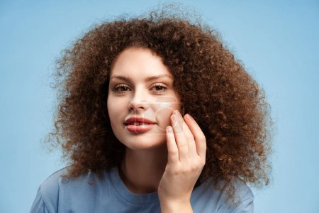Téléchargez les photos : Smiling beautiful young woman with curly hair applying cream on face, looking in mirror, standing, isolated on blue background. Concept of skin care, moisturizing, hydration - en image libre de droit