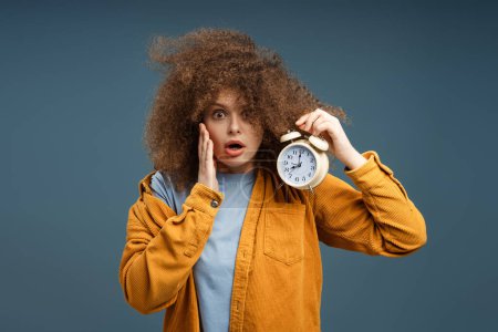 Photo for Amazed, shocked young woman with curly hair holding alarm clock, late, looking at camera, standing isolated on black background. Concept of morning, sleep - Royalty Free Image