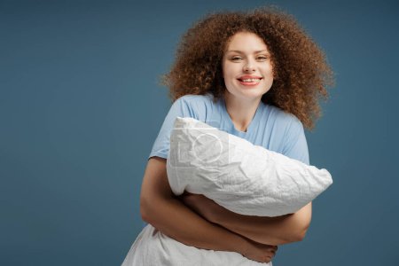 Photo for Cheerful beautiful woman with curly hair holding pillow, smiling, looking at camera, standing isolated on blue background. Concept of advertisement, shopping - Royalty Free Image