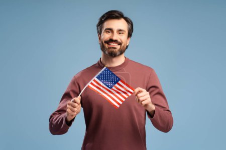 Téléchargez les photos : Portrait d'un homme séduisant regardant la caméra et tenant le drapeau national des États-Unis, isolé sur fond bleu. Fête du Souvenir, fête de l'indépendance. - en image libre de droit