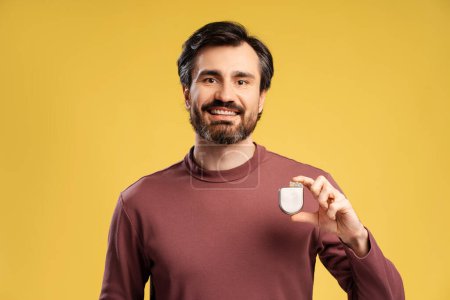 Handsome smiling bearded man holding cardiac pacemaker looking at camera, isolated on yellow. Health care, treatment concept