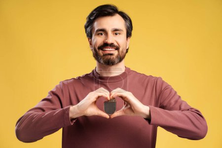 Portrait of happy bearded man holding cardiac pacemaker in two hands, looking at camera, isolated on yellow. Health care, treatment concept