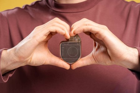 Cropped photo of male hands holding cardiac pacemaker, looking at camera, isolated on yellow background. Health care, treatment concept