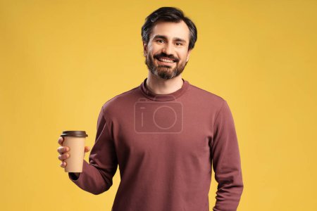 Photo for Portrait of smiling bearded man holding paper cup of coffee while posing in studio, looking at camera, isolated on yellow background - Royalty Free Image
