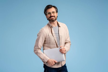 Glücklicher bärtiger Mann mit Brille, Laptop in die Kamera blickend, isoliert auf blauem Hintergrund stehend. Bildung, Geschäftskonzept, Online-Technologie